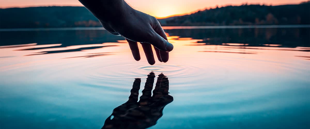 A hand touching the surface of a calm lake at sunset